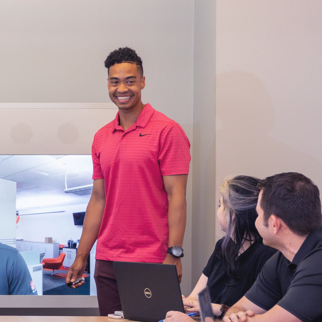 Male Store Support Center associate in pink polo shirt leads a meeting in a boardroom