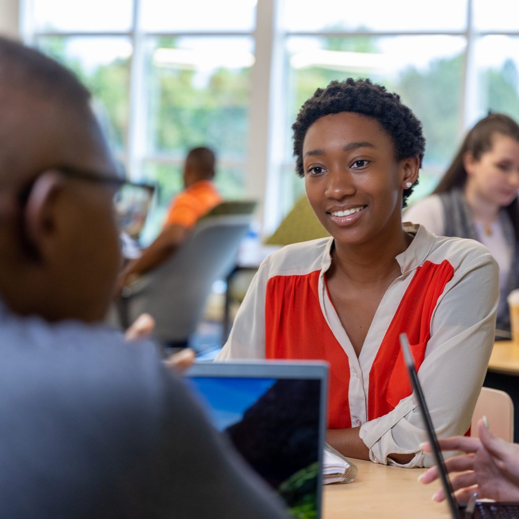 Female Store Support Center listens to coworkers while sitting at a table in a common area